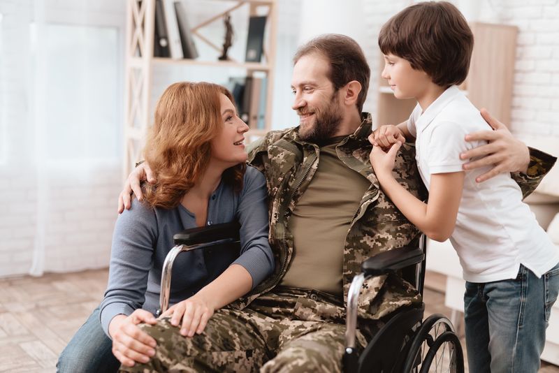 A veteran in uniform sits in a wheelchair at home while his wife and son hug him.